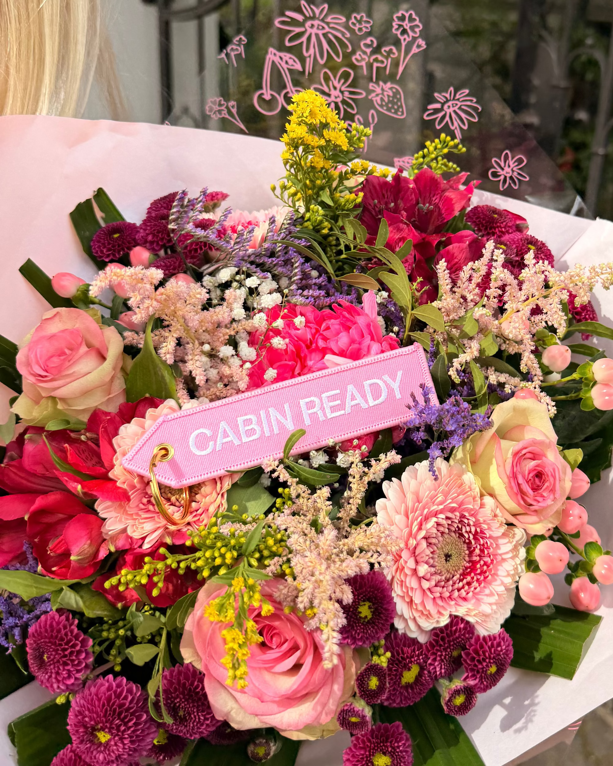 Rosa „Cabin Ready“ Schlüsselanhänger mit weißer Stickerei und goldfarbenem Ring vor buntem Blumenstrauss