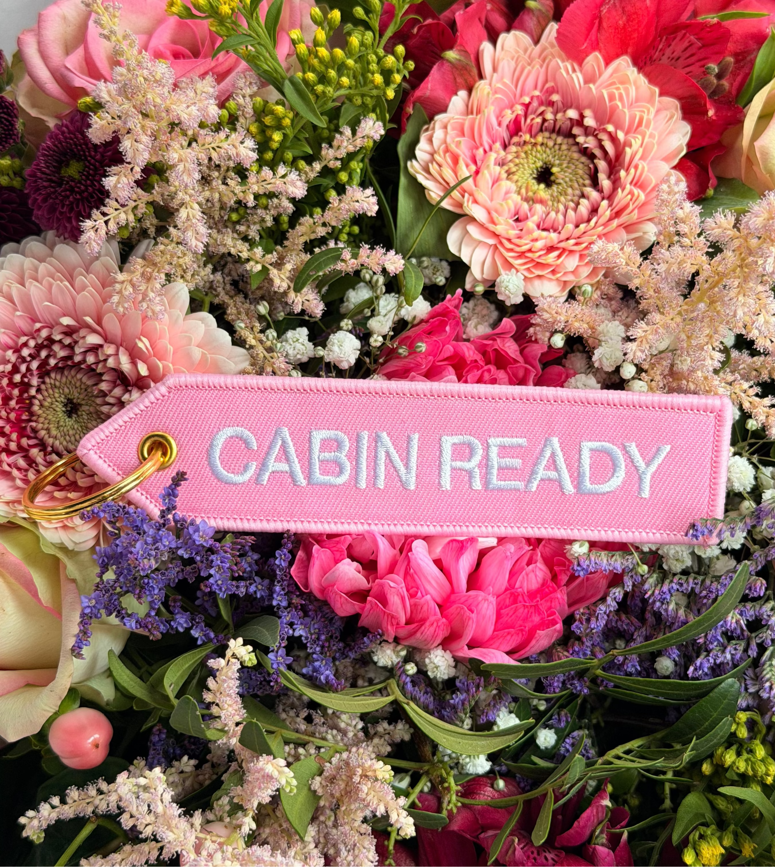 Rosa „Cabin Ready“ Schlüsselanhänger mit weißer Stickerei und goldfarbenem Ring vor Blumen